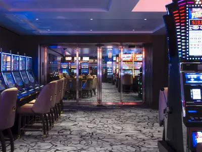 Eine weite Aufnahme des Casinos an Bord der Norwegian Prima. Reihen von leuchtenden Spielautomaten säumen den Raum, gespiegelt in den Glastüren zur Casino-Lounge. Elegante Barhocker laden zum Verweilen ein.