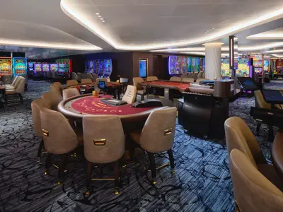 Blick in das moderne Casino der Norwegian Prima mit zahlreichen, leuchtenden Slot-Maschinen und eleganten Spieltischen. Mehrere Tische mit Stuehlen sind im Vordergrund zu sehen.