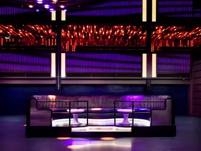 Moderner Club-Lounge-Bereich an Bord der Norwegian Prima. Eine geschwungene, violett beleuchtete Sitzbank mit Tischen und roten Deckenlichtern. Keine Personen.