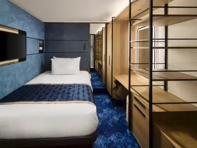 Eine stilvolle Einzelkabine an Bord der Norwegian Prima mit einem Einzelbett, Flachbildfernseher, blauem Teppich und Holzmöbeln. Die Kabine ist in Blau- und Holztönen gehalten und modern eingerichtet.
