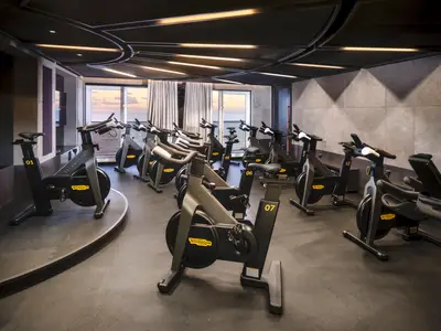 Blick in das moderne Spinning-Studio an Bord der Norwegian Prima. Mehrere schwarze Technogym-Fahrräder stehen bereit, mit großen Fenstern zum Meer, das im Sonnenuntergang leuchtet.