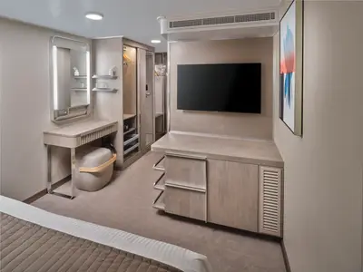Blick in eine moderne Innenkabine an Bord der Norwegian Prima. Zu sehen sind ein heller Holztisch mit Spiegel, ein Hocker, ein großer Fernseher und ein offener Kleiderschrank mit Ablagen.