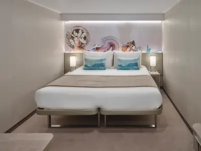 Moderne Innenkabine an Bord der Norwegian Prima. Ein Doppelbett mit blauen Kissen und beleuchteter Wandkunst sind zentral. Nachttische mit Lampen stehen seitlich.
