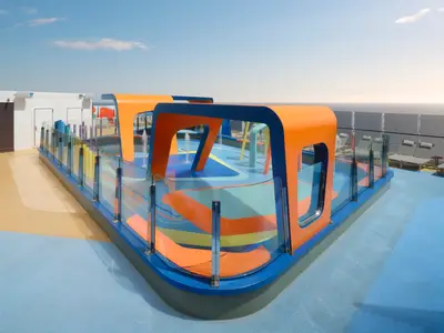 Ein farbenfroher Kinder-Aqua-Park auf dem Oberdeck der Norwegian Prima mit orangen, blauen und aquafarbenen Spielelementen. Mehrere kleine Wasserfontänen spritzen aus dem Boden, im Hintergrund ist das weite Meer unter blauem Himmel zu sehen. Die Anlage ist von einer Glasbrüstung umgeben.
