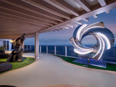 Ein Aussendeck der Norwegian Prima bei Dämmerung mit zwei modernen Skulpturen. Links eine dunkle, geometrische Wolfsstatue, rechts eine leuchtende, spiralförmige Metallskulptur vor dem Meer.