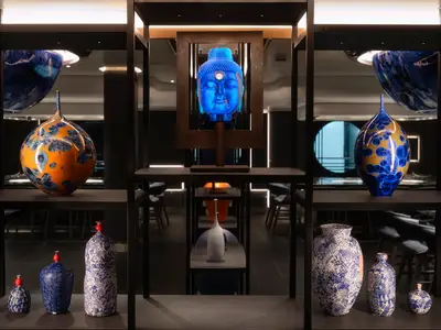 Kunstobjekte auf schwarzen Regalen an Bord der Norwegian Prima. Zentral ein leuchtend blauer Buddha-Kopf, flankiert von großen, bunten Vasen mit kristallinen Mustern und kleineren blau-weißen Porzellanvasen.