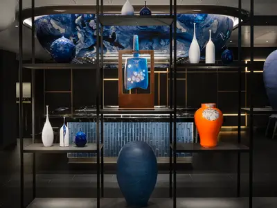 Eine moderne Kunstinstallation an Bord der Norwegian Prima zeigt blaue und weisse Keramikvasen sowie eine grosse blaue Glasskulptur in einem Metallrahmen. Die Exponate stehen auf dunklen Regalen vor einem Hintergrund aus dunkelblauen Fliesen und goldenen Akzenten.