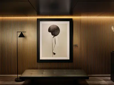 Ein eleganter Flur an Bord der Norwegian Prima. Die Wände sind mit vertikalen Holzlatten verkleidet, die von indirektem Licht beleuchtet werden. Eine gerahmte Schwarz-Weiß-Fotografie einer Person mit muschelartigem Kopfschmuck hängt mittig. Rechts davon steht eine goldene, abstrakte Skulptur auf einem dunklen Podest. Eine schwarze Sitzbank und eine Stehlampe ergänzen die minimalistische Einrichtung.