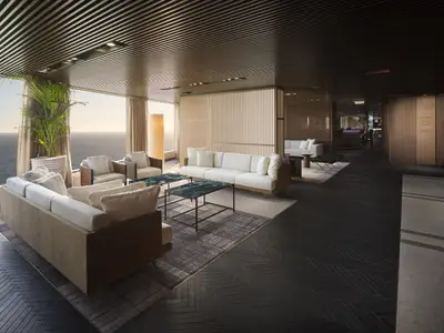 Elegante Haven Lounge der Norwegian Prima. Moderne, helle Sofas und Sessel, dunkle Tische auf Teppich und Fischgrätboden. Panoramablick auf das Meer bei Sonnenuntergang.