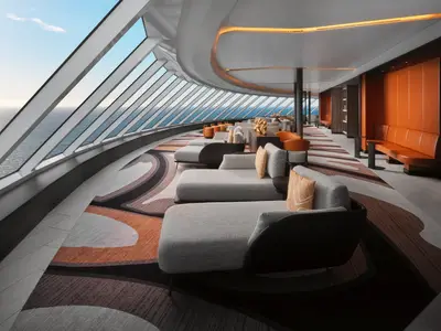 Blick in die elegante Observation Lounge an Bord der Norwegian Prima. Zahlreiche bequeme Loungemöbel stehen entlang der großen Fensterfront, die einen weiten Blick auf das offene Meer bietet. Die Lounge ist in warmen Orange- und Brauntönen gehalten.