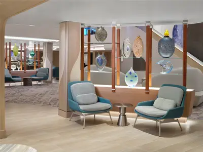 Ein heller, eleganter Atrium-Bereich an Bord der Norwegian Prima. Türkisblaue Sessel umgeben einen Beistelltisch, während kunstvolle, farbige Glasvasen auf beleuchteten Regalen ausgestellt sind.