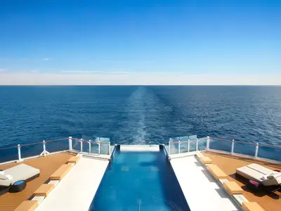 Ein Infinity-Pool am Heck der Norwegian Prima bei Tag, umgeben von Holzdecks und Liegestühlen. Der Pool bietet einen nahtlosen Blick auf das offene, blaue Meer und die weisse Heckwelle des Schiffes.