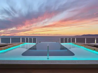 Ein Infinity-Pool mit leuchtend blauem Wasser und Liegestühlen an Deck der Norwegian Prima bei einem dramatischen Sonnenuntergang über dem Meer und fernen Inseln.
