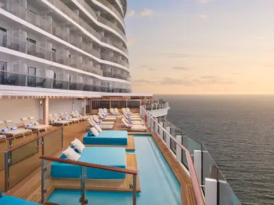 Das Sonnendeck der Norwegian Prima mit Infinity-Pool, blauen Liegeflaechen und Sonnenliegen bei Sonnenuntergang. Im Hintergrund die Balkonkabinen und das weite Meer.