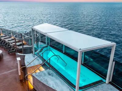 Blick von oben auf den beleuchteten Infinity-Pool mit türkisblauem Wasser und Whirlpool-Funktion an Bord der Norwegian Prima. Rechts des Pools sind Sonnenliegen und ein gläsernes Geländer zu sehen, das einen Panoramablick auf das Meer freigibt.