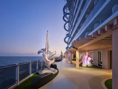 Abendstimmung auf der Norwegian Prima mit einem modernen Skulpturengarten auf dem Außendeck, Blick auf das Meer und blaue Wasserrutschen an der Schiffsseite.