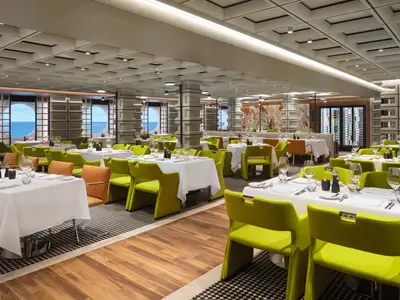 Modernes Spezialitätenrestaurant an Bord der Norwegian Prima. Viele weiß gedeckte Tische mit grünen Stühlen. Runde Fenster bieten Meerblick; Weinwand und Kunst im Hintergrund.