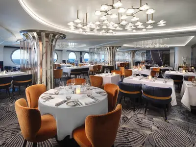Blick in das elegante Restaurant 'The Commodore Room' an Bord der Norwegian Prima. Zahlreiche weiß gedeckte Tische mit blauen und orangefarbenen Stühlen. Große runde Fenster bieten Meerblick.