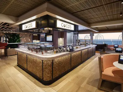 Das helle und moderne Coco's Dessert-Café an Bord der Norwegian Prima zeigt eine lange Marmortheke voller Süßspeisen und bunter Bonbons, gemütliche Sitzecken und einen weiten Meerblick durch große Fenster.