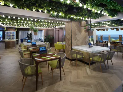 Restaurantbereich Indulge Food Hall an Bord der Norwegian Prima. Viele Tische mit hellgrünen Stühlen, Deckenbegrünung, Lichterketten und weitem Blick auf das Meer. Ohne Personen.