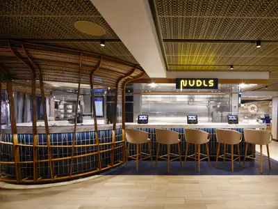 Ansicht des modernen NUDLS Restaurants an Bord der Norwegian Prima. Ein Tresen mit blauen Fliesen und hellen Hockern. Über dem Tresen leuchtet der NUDLS-Schriftzug. Die Decke ist golden gemustert.