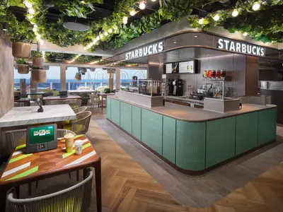 Starbucks Café an Bord der Norwegian Prima mit langer grüner Theke. Sitzbereiche mit Meerblick. Auf einem Tisch stehen Tablet und zwei Starbucks-Getränke. Decke mit Pflanzen und Lichterketten ziert den Raum.