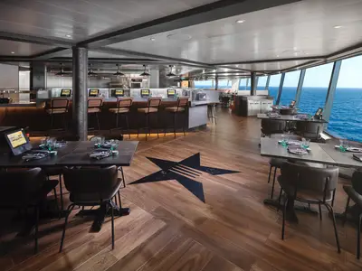Innenansicht des modernen Food Republic Restaurants an Bord der Norwegian Prima, mit Tischen, Barhockern, offener Küche und großen Panoramafenstern zum Meer.