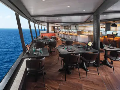 Innenansicht des modernen Spezialitätenrestaurants Food Republic an Bord der Norwegian Prima, mit Tischen, Stühlen, einer Bar und großen Panoramafenstern mit Blick auf das offene Meer.