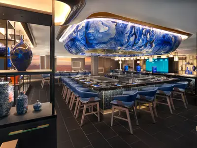Das moderne Hasuki Teppanyaki Restaurant an Bord der Norwegian Prima. Dunkle Tische mit Kochplatten, blaue Barhocker, kunstvolle blau-weiße Decke und Fenster mit Meerblick.