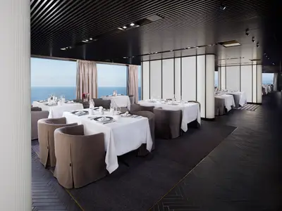 Ein elegantes Restaurant an Bord der Norwegian Prima mit Tischen, die mit weißen Tischdecken, Besteck und Gläsern gedeckt sind. Große Fenster bieten einen weiten Blick auf das offene Meer. Moderne Beleuchtung und Inneneinrichtung.