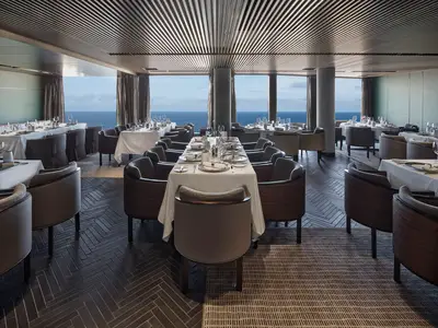 Innenansicht eines modernen, eleganten Restaurants mit dunklem Holzfussboden und grossen Panoramafenstern mit Blick auf das offene Meer, an Bord der Norwegian Prima. Viele Tische sind weiss gedeckt und bereit für Gäste.