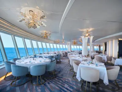 Blick in das elegant gedeckte Restaurant Hudson's an Bord der Norwegian Prima mit Tischen und Stühlen. Große Fensterfronten bieten einen weiten Meerblick. Goldene Designer-Kronleuchter zieren die Decke.