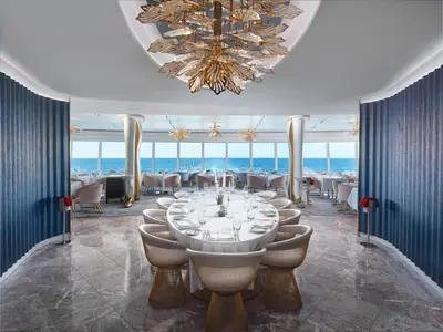 Ein helles, ovales Restaurant an Bord der Norwegian Prima mit grossen Panoramafenstern zum Meer. Die Tische sind weiss gedeckt, die Stühle hell. Eine goldene Deckenleuchte und blauen Wandpaneele sind sichtbar.