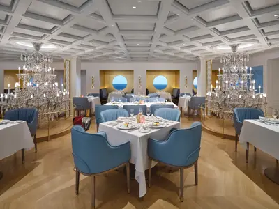 Elegantes Restaurant Le Bistro an Bord der Norwegian Prima. Man sieht blau gepolsterte Stühle, weiß gedeckte Tische, große Kronleuchter und runde Fenster mit Meerblick.