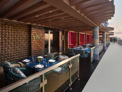 Blick auf das Außenrestaurant Los Lobos an Bord der Norwegian Prima bei Sonnenuntergang. Blaue Tische, gemusterte Stühle und ein leuchtendes Logo sind zu sehen, dahinter das Meer.