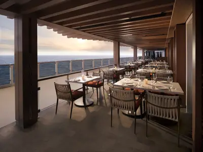 Elegantes Außendeck-Restaurant an Bord der Norwegian Prima. Zahlreiche Tische und Stühle unter einer Holzlamellen-Überdachung. Freier Blick auf das weite Meer beim Sonnenuntergang.