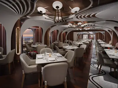 Elegantes Spezialitätenrestaurant der Norwegian Prima mit beigen Stühlen, modernen Lichtinstallationen und Meerblick bei Sonnenuntergang.
