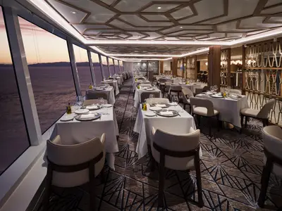 Das elegante Spezialitätenrestaurant Palomar an Bord der Norwegian Prima mit gedeckten Tischen entlang großer Fenster. Meerblick bei Sonnenuntergang. Stilvolles Interieur mit gemusterter Decke und Teppich. Ohne Personen.