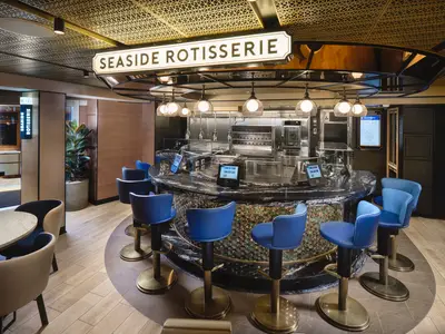 Ansicht der Seaside Rotisserie an Bord der Norwegian Prima. Ein eleganter Raum mit offener Kueche, einer Theke aus dunklem Marmor und blauen Barhockern mit goldfarbenen Fuessen.