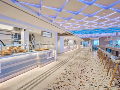 Ein helles, geräumiges Buffetrestaurant Surfside Cafe an Bord der Norwegian Prima. Es gibt lange Essensausgaben mit Gebäck und Speisen, sowie eine Bar mit Hockern und Meerblick durch große Fenster. Die Decke ist mit blauer und weißer LED-Beleuchtung modern gestaltet.
