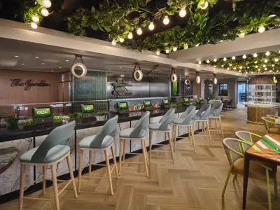 Blick in das Restaurant 'The Garden' an Bord der Norwegian Prima. Eine moderne Selbstbedienungstheke mit frischen Zutaten und Bestell-Terminals ist sichtbar. Zahlreiche Barhocker stehen davor. Die Decke ist mit Pflanzen und Lichterketten dekoriert.