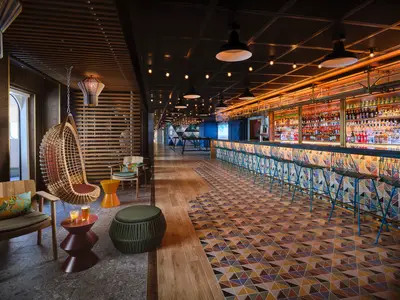 Moderne Bar 'The Local' auf der Norwegian Prima. Eine lange Theke mit bunten Barhockern. Links ein gemütlicher Loungebereich mit einem Hängesessel und bunten Hockern.