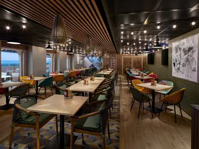 Blick in das legere Restaurant "The Local" an Bord der Norwegian Prima mit Tischen, bunten Stühlen und großen Fenstern zum Meer. Die Decke ist mit Holzlamellen und Lichterketten gestaltet.
