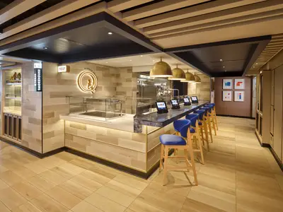 Innenansicht des modernen Q Texas Smokehouse an Bord der Norwegian Prima mit langer Bar, blauen Hockern und beleuchtetem Logo.
