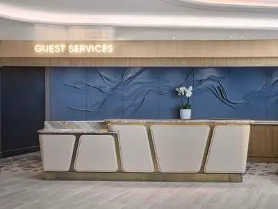 Moderne Rezeption an Bord der Norwegian Prima. Ein beleuchteter 'Guest Services' Schriftzug ist über einer hellen Theke mit Marmorplatte zu sehen. Die blaue Rückwand ist mit einem Wellenrelief und einer weißen Orchidee gestaltet.