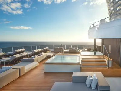 Das Sonnendeck der Norwegian Prima zeigt zwei Whirlpools, moderne Lounge-Möbel und Liegen. Im Hintergrund ist das Meer unter einem klaren Himmel bei Sonnenuntergang zu sehen.
