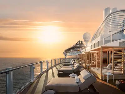 Blick über das Sonnendeck der Norwegian Prima bei Sonnenuntergang. Zahlreiche leere Liegestühle und Holz-Cabanas reihen sich auf dem Deck entlang des Ozeans, dessen Oberfläche im goldenen Licht glänzt. Die Sonne steht tief am Horizont, taucht Himmel und Meer in warme Orange- und Gelbtöne und reflektiert auf den gläsernen Begrenzungen des Decks.