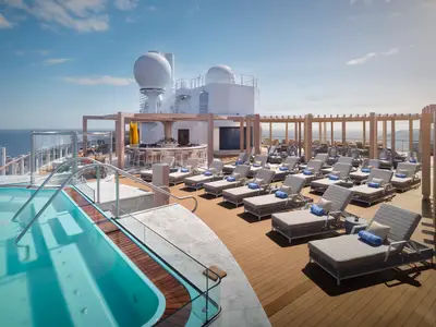 Das sonnige Sonnendeck der Norwegian Prima. Im Vordergrund ein Pool, dahinter eine Bar und viele Liegen mit blauen Handtüchern. Im Hintergrund das weite Meer unter blauem Himmel.