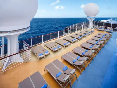 Ein weitläufiges Sonnendeck der Norwegian Prima mit Reihen von braunen Sonnenliegen, blauen Handtüchern und Kissen. Im Hintergrund erstreckt sich das weite blaue Meer unter einem hellen Himmel. Die Szene ist von großen weißen Aufbauten eingerahmt.