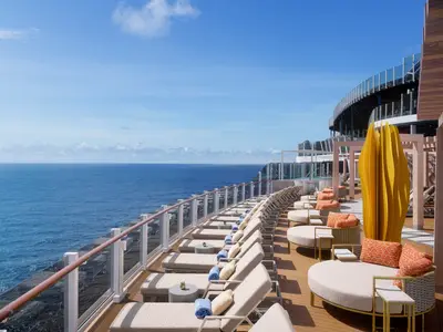 Sonnendeck der Norwegian Prima mit zahlreichen hellen Sonnenliegen und Lounge-Bereichen. Im Vordergrund eine gelbe Skulptur. Rechts ein oberes Deck und links der weite Ozean unter blauem Himmel.